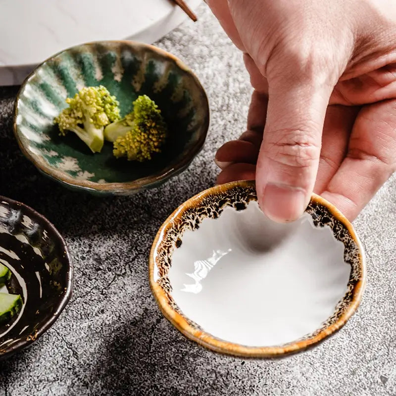 Une main tient un petit bol à condiments en céramique blanche, avec une bordure brune mouchetée et un motif de glaçure peint à la main, d’une grande finesse.