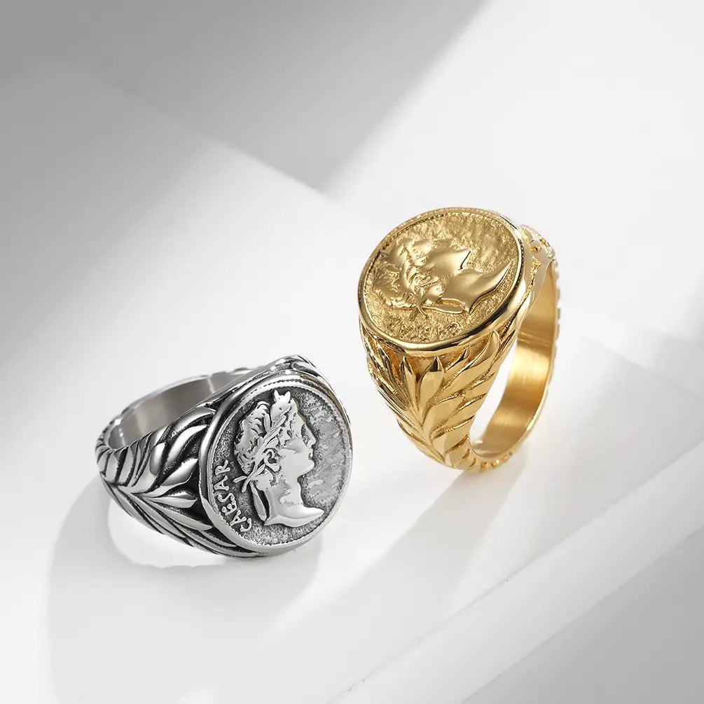 Two vintage Caesar portrait stainless steel signet rings, one silver with a detailed profile portrait and leafy band design, and one gold with an embossed face on its circular face.