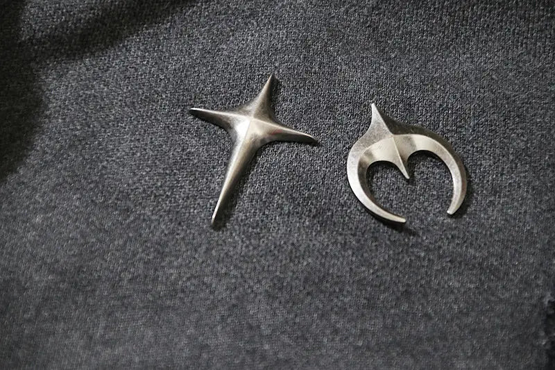 Dos broches distintivos con acabado plateado metálico con forma de estrella de cinco puntas y una media luna abstracta con un centro en forma de corazón, apoyados sobre la tela gris oscuro de un cárdigan con cremallera y capucha estilo batik desgastado.
