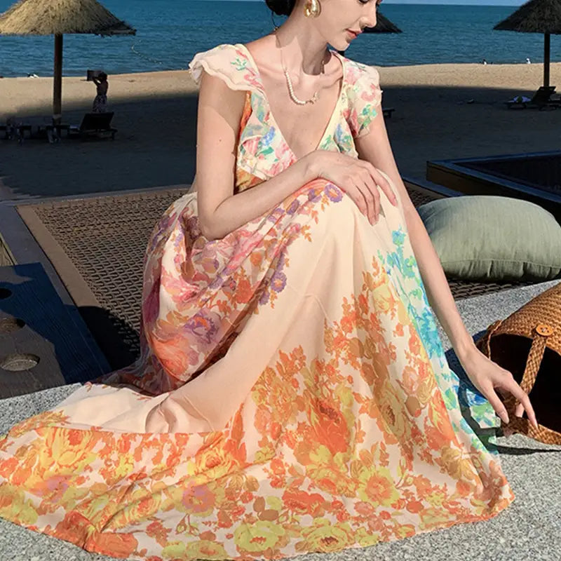 A flowing sleeveless french floral v neck ruffle maxi dress in a soft peach base with vibrant orange and yellow floral patterns.
