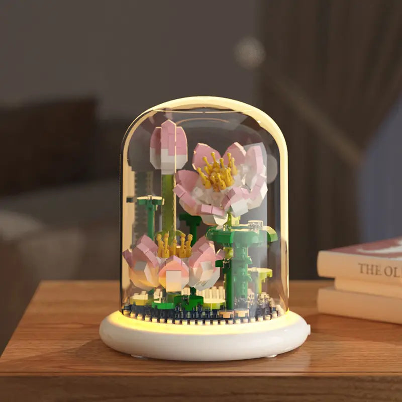 Une délicate sculpture de fleur de lotus rose et blanche en briques plastiques emboîtées, exposée sous une coupole en verre transparente, avec une base doucement lumineuse, comme décoration jouet de dôme en verre pour fleurs, bloc de construction, éclairage LED.