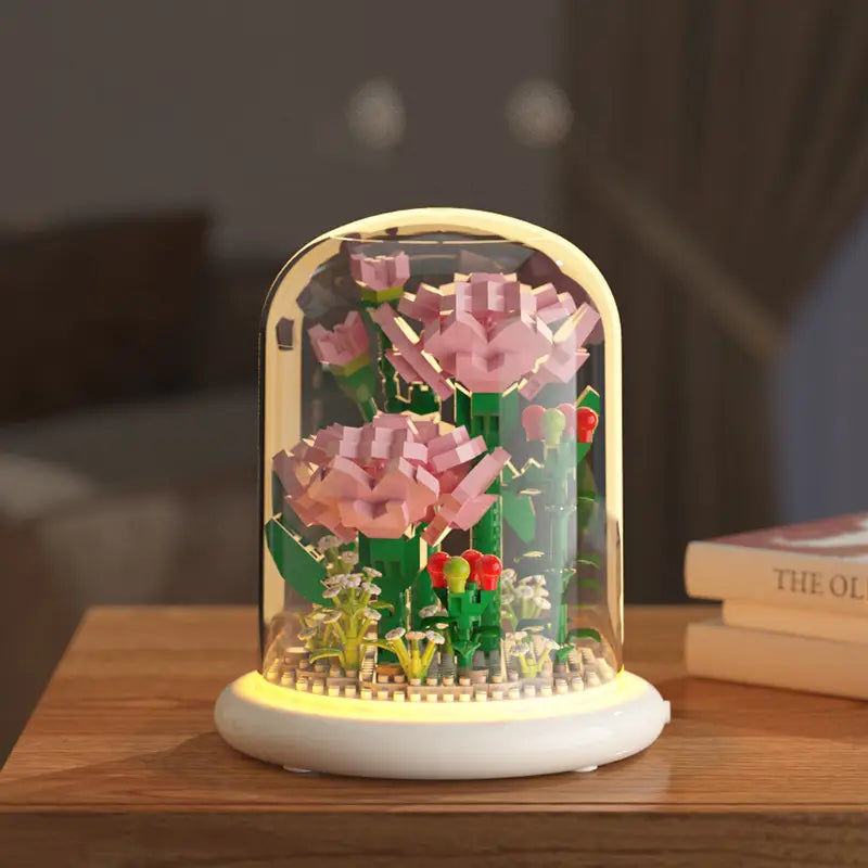 Un présentoir floral style LEGO rose et vert fabriqué en éléments de construction, sous une coupole en verre transparente, avec une base illuminée, comme décoration jouet de dôme en verre pour fleurs, bloc de construction.
