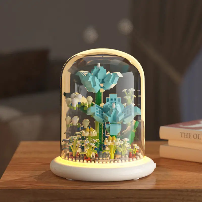 Un jouet décoratif de dôme en verre pour fleurs, bloc de construction, éclairage LED, présentant une miniature de jardin de style LEGO, avec des fleurs en blocs turquoise et vertes sous un éclairage intérieur chaud.