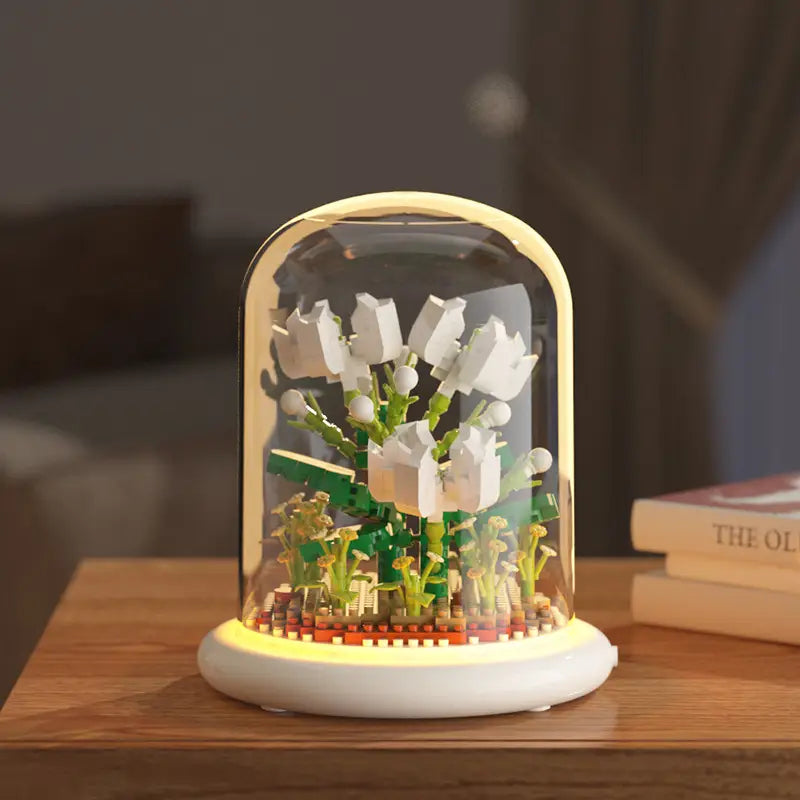 Un modèle LEGO blanc et vert de muguet exposé à l’intérieur d’une coupole en verre transparente, avec un éclairage LED chaud, comme décoration jouet de dôme en verre pour fleurs, bloc de construction.