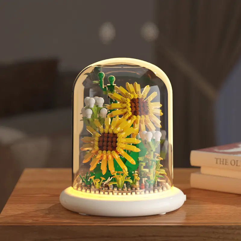 Un jouet décoratif de dôme en verre pour fleurs, bloc de construction, éclairage LED, présentant deux tournesols jaunes vibrants avec des centres bruns, entourés d’un feuillage vert et de fleurs blanches sous une coupole en verre transparente.