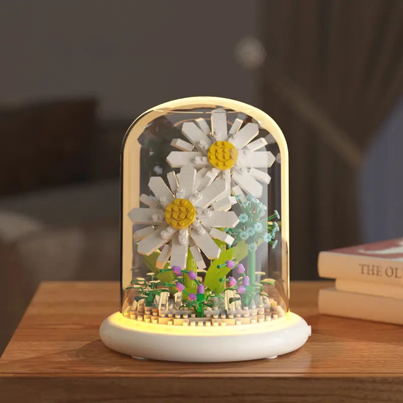 Un présentoir de dôme de fleurs LEGO blanc et jaune, avec deux marguerites à tiges vertes et des fleurs violettes, illuminées par une base lumineuse à LED, comme décoration jouet de dôme en verre pour fleurs, bloc de construction.