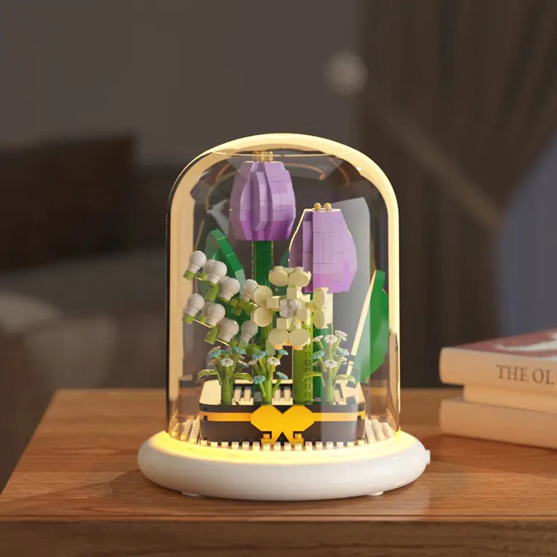 Un présentoir de fleurs LEGO violet et blanc sous une coupole en verre transparente, avec une base à LED lumineuse conçue comme jouet décoratif en forme de bloc de construction.