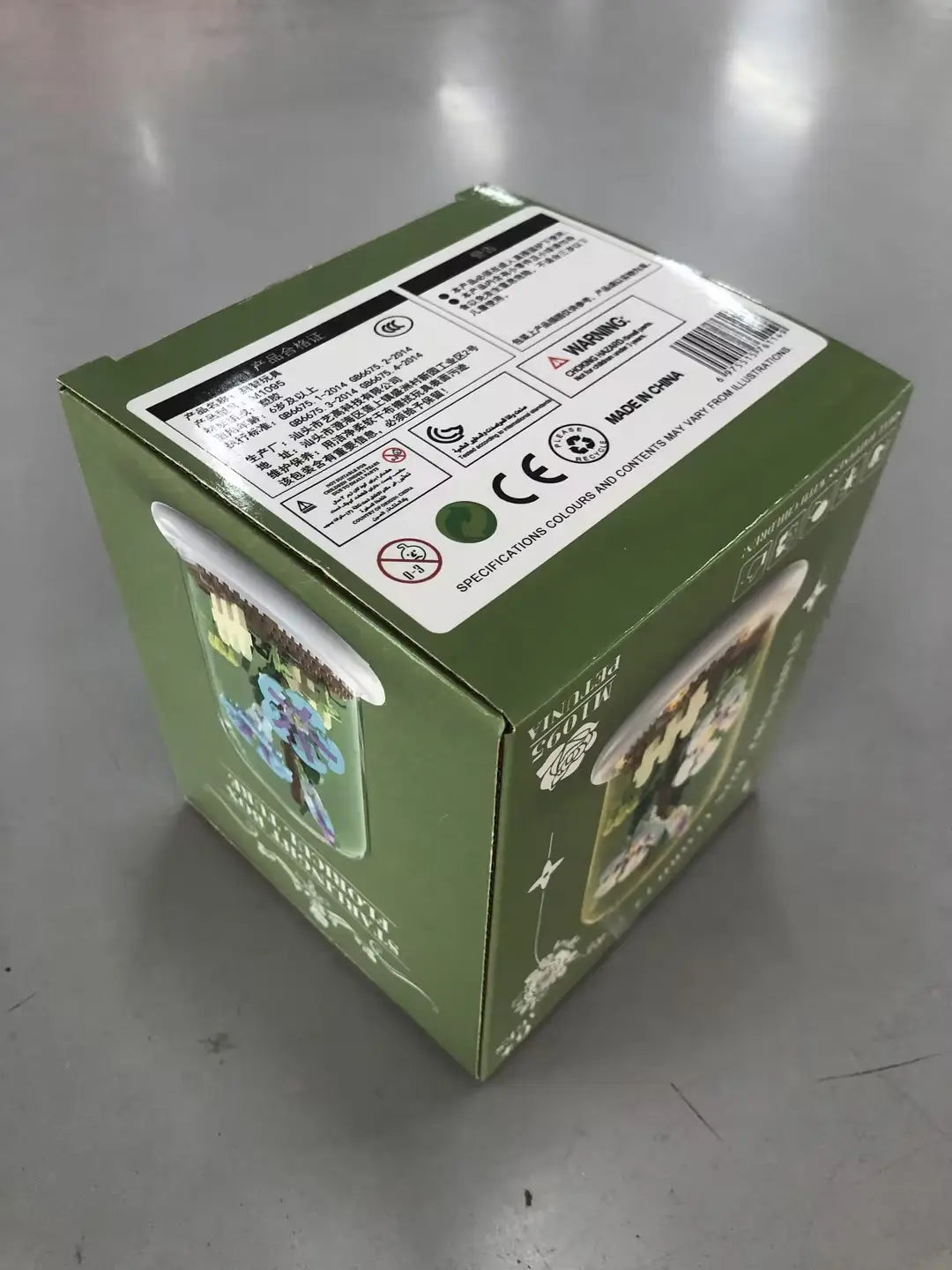 Boîte en carton verte avec des illustrations imprimées d’une scène fantastique et des étiquettes réglementaires pour l’emballage du jouet décoratif de dôme en verre pour fleurs, bloc de construction, éclairage LED.