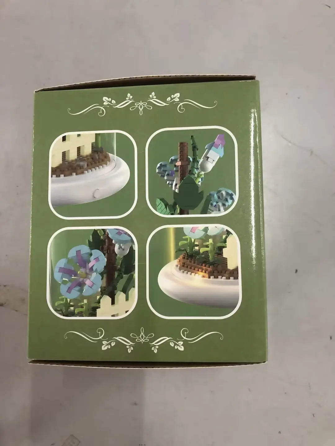 Boîte en carton verte pour le jouet décoratif de dôme en verre pour fleurs, bloc de construction, éclairage LED, présentant quatre images circulaires de scènes de jardin miniatures de style LEGO, avec des plantes et des fleurs colorées, ainsi qu’une base blanche.