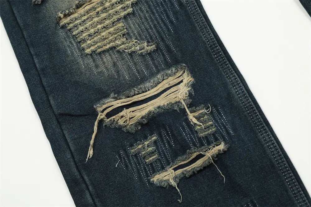 Jeans déchirés homme à effet usé, jambe droite, en denim bleu foncé, comportant plusieurs zones de déchirure effilochées avec coutures visibles et bords bruts.