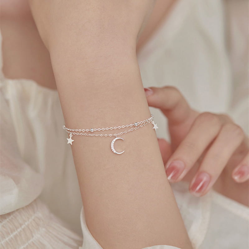 A delicate silver double-layered anklet bracelet featuring a crescent moon charm and small star charms adorned with tiny sparkling stones.