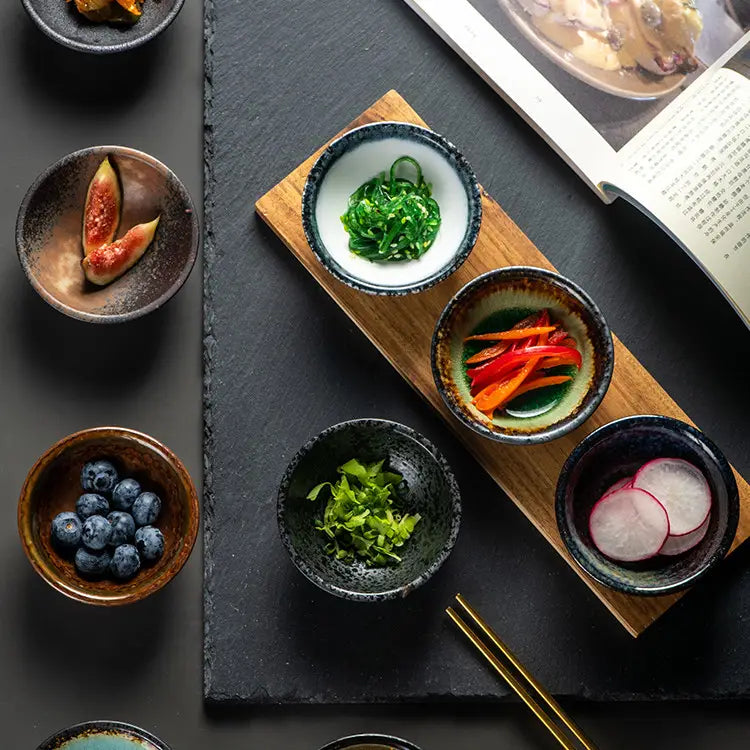 Set of five Japanese ceramic small plates with rustic speckled glaze each holding different colorful garnishes.