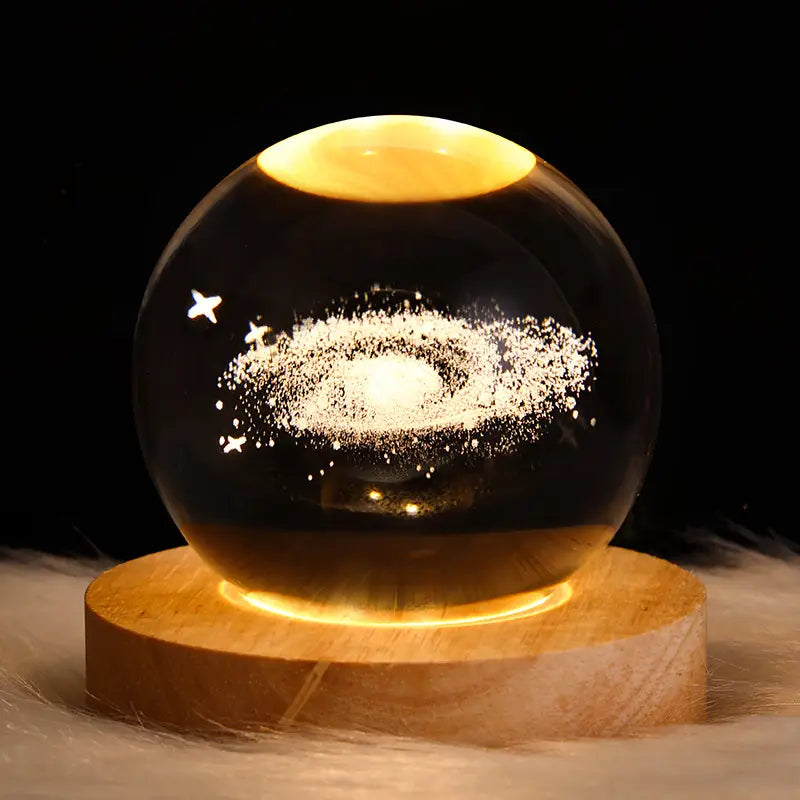 A glowing 3d galaxy crystal ball led night light moon lamp features a glass orb with a galaxy design of white stars and swirling cosmic dust illuminated by warm yellow light from within.