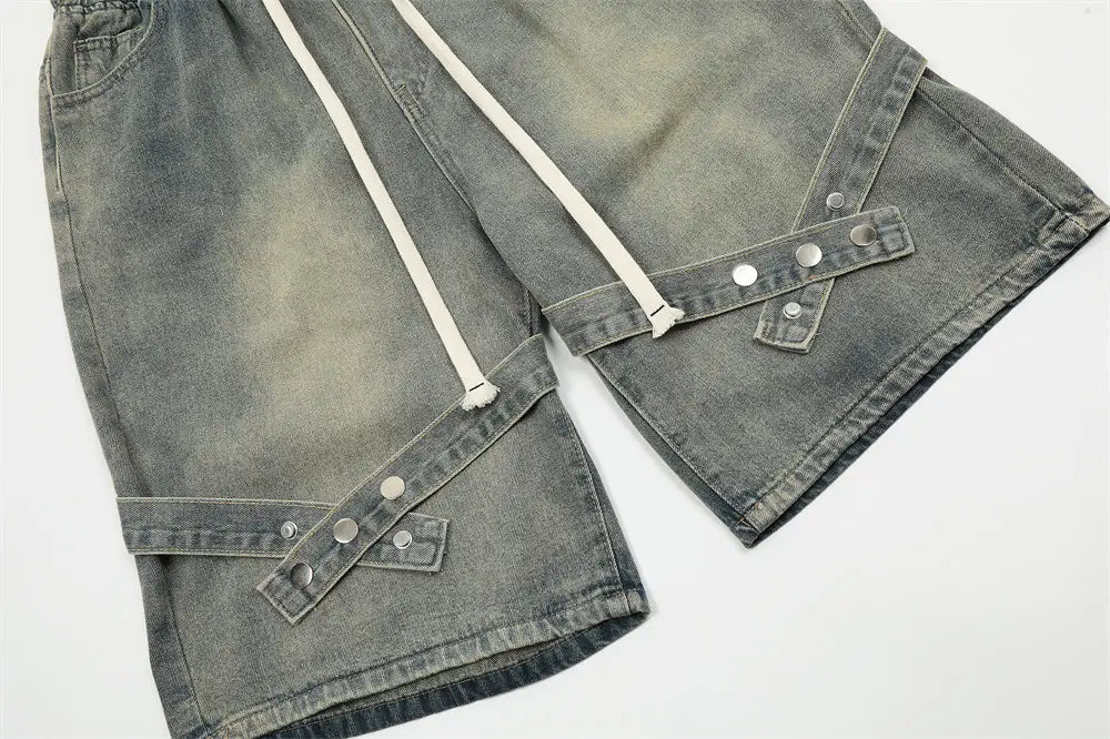 Shorts en jean gris à bretelles diagonales pour hommes, avec taille à cordon blanc et accents de boutons argentés.