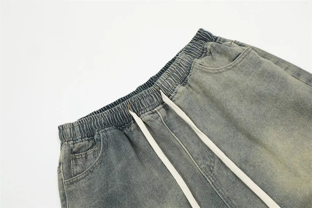 Joggers en jean gris pour hommes, coupe décontractée, dotés d’une ceinture élastique et de cordons blancs contrastants, complétant le design à bretelle diagonale des shorts en jean à taille à cordon.