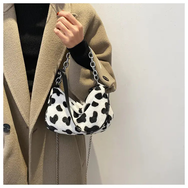 A plush black and white cow print crossbody baguette bag with a silver chain strap.