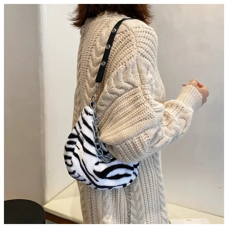 A plush zebra-striped baguette bag made of soft material with a black strap and silver chain accents.