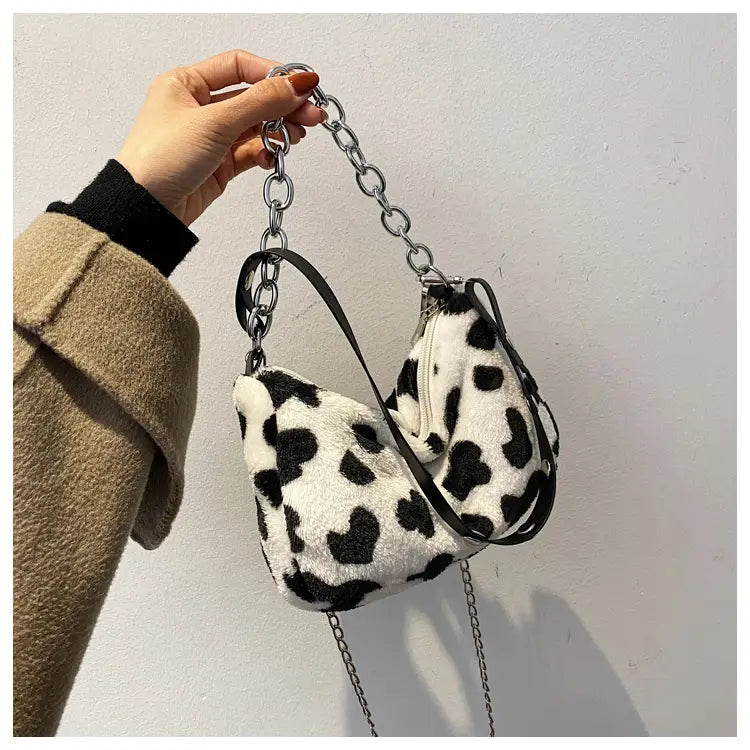 A cozy black-and-white cow print faux fur crossbody baguette bag with a silver chain strap and black leather accents.