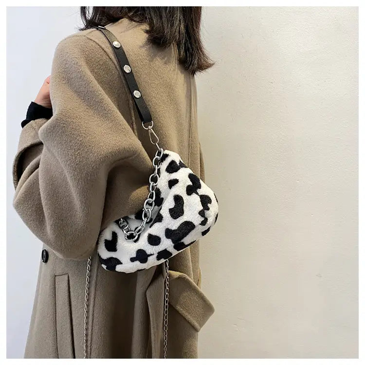 A plush white and black leopard-print crossbody baguette bag made of soft fabric with a silver chain strap.