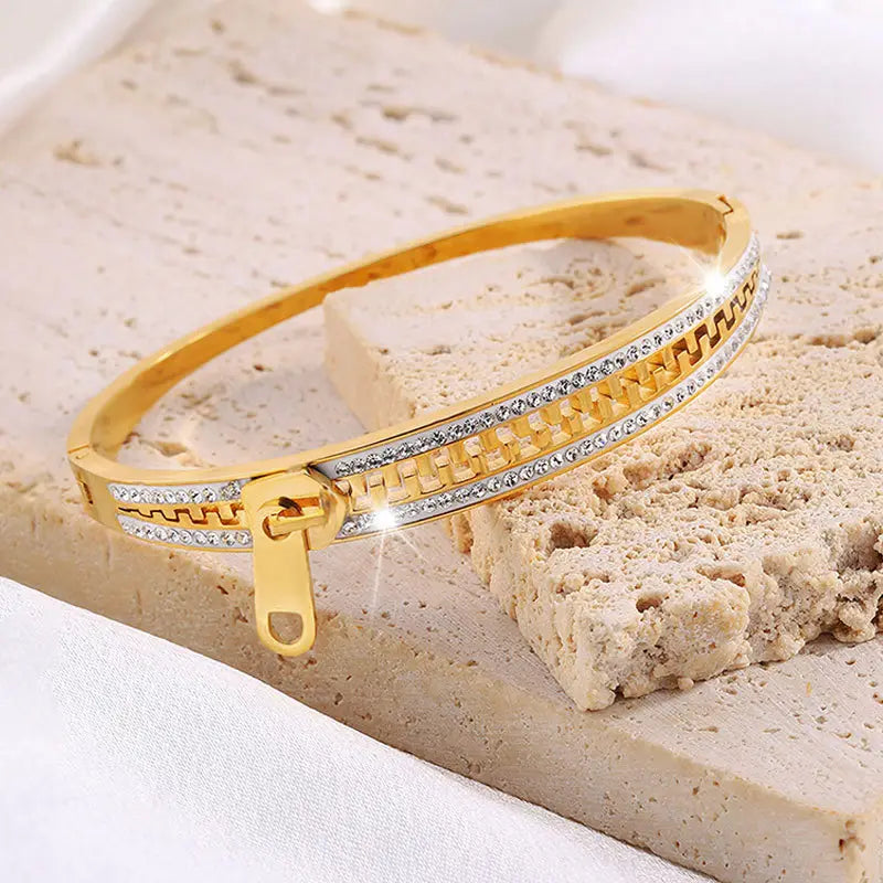 Gold-toned crystal interlocking titanium steel zipper bangle bracelet with embedded clear crystals along its textured band and zipper closure.