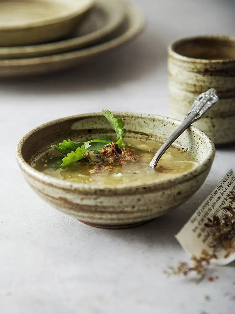 Earthenware bowl from ceramic rustic speckled stoneware tableware set with speckled beige glaze and horizontal ridges holding light-colored soup garnished with fresh cilantro.