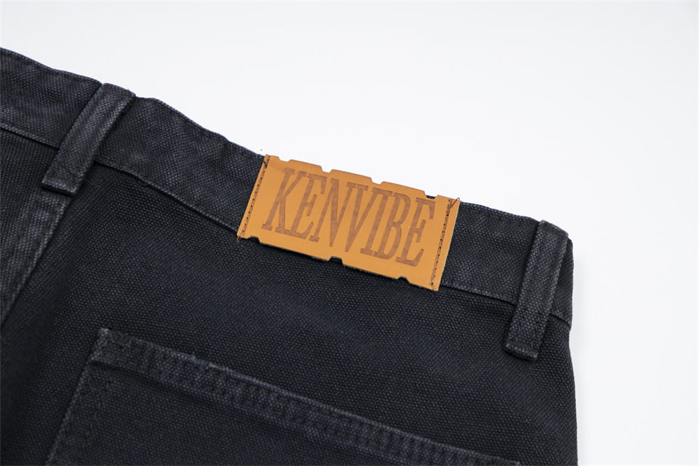 Pantalones cortos de mezclilla negros desgastados para hombre con dobladillo deshilachado, con un parche de cuero color camel grabado con el nombre de la marca kenvibe en la parte posterior.