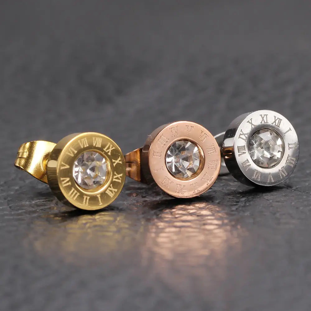 Trois élégantes boucles d’oreilles à tige dans des finitions or, or rose et argent, comprenant des formes rondes avec des chiffres romains et des cristaux transparents centraux, issues de la parure de boucles d’oreilles à tige en diamant en or rose, motif de chiffres romains.