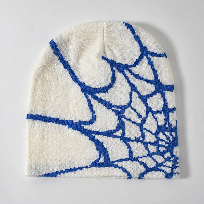 A cream-colored knit beanie winter hat featuring a bold blue spiderweb pattern woven into the fabric.