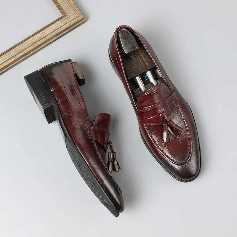Mocasines con borlas de cuero con estampado de cocodrilo color burdeos con acabado pulido y suelas negras de goma, mostrados desde la parte trasera.