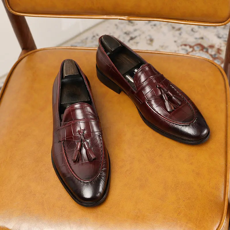 Un par de mocasines con borlas de cuero con estampado de cocodrilo color burdeos que muestran su rica textura y diseño elegante.