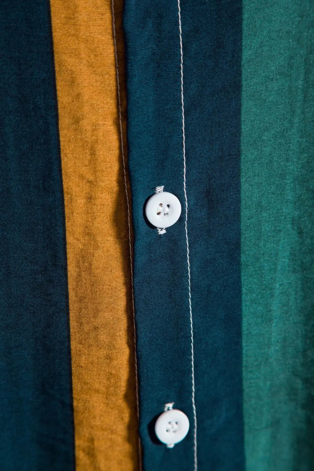 A white four-hole button sewn onto a striped short sleeve button up shirt with deep teal and mustard yellow vertical stripes.