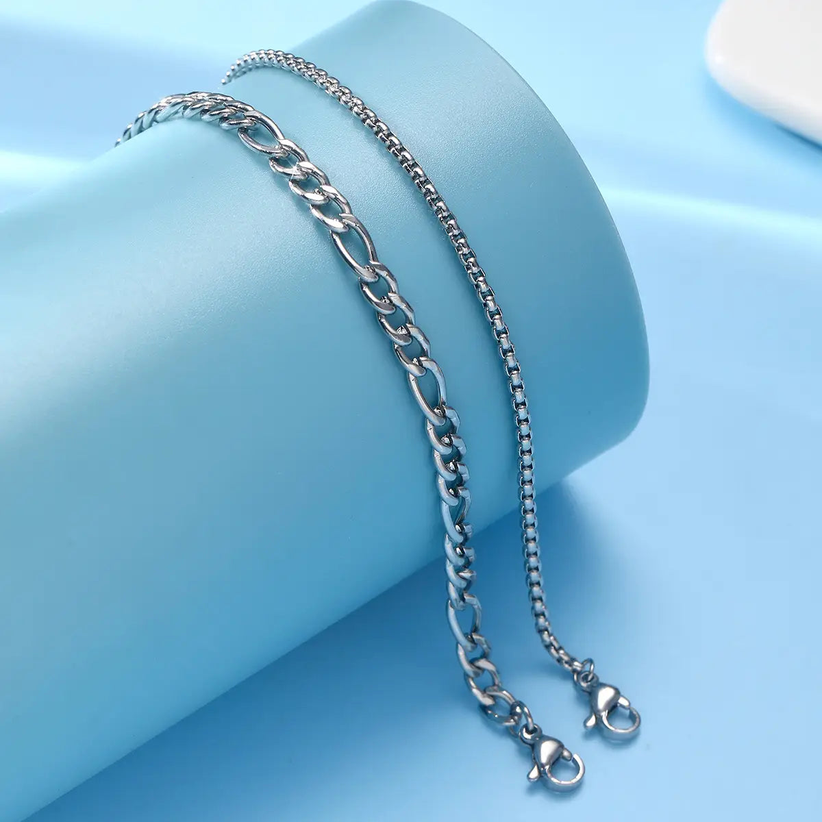 Two silver-toned unisex bracelets featuring a bold figaro link design and a delicate box chain pattern from the layered Cuban and figaro link bracelet set.