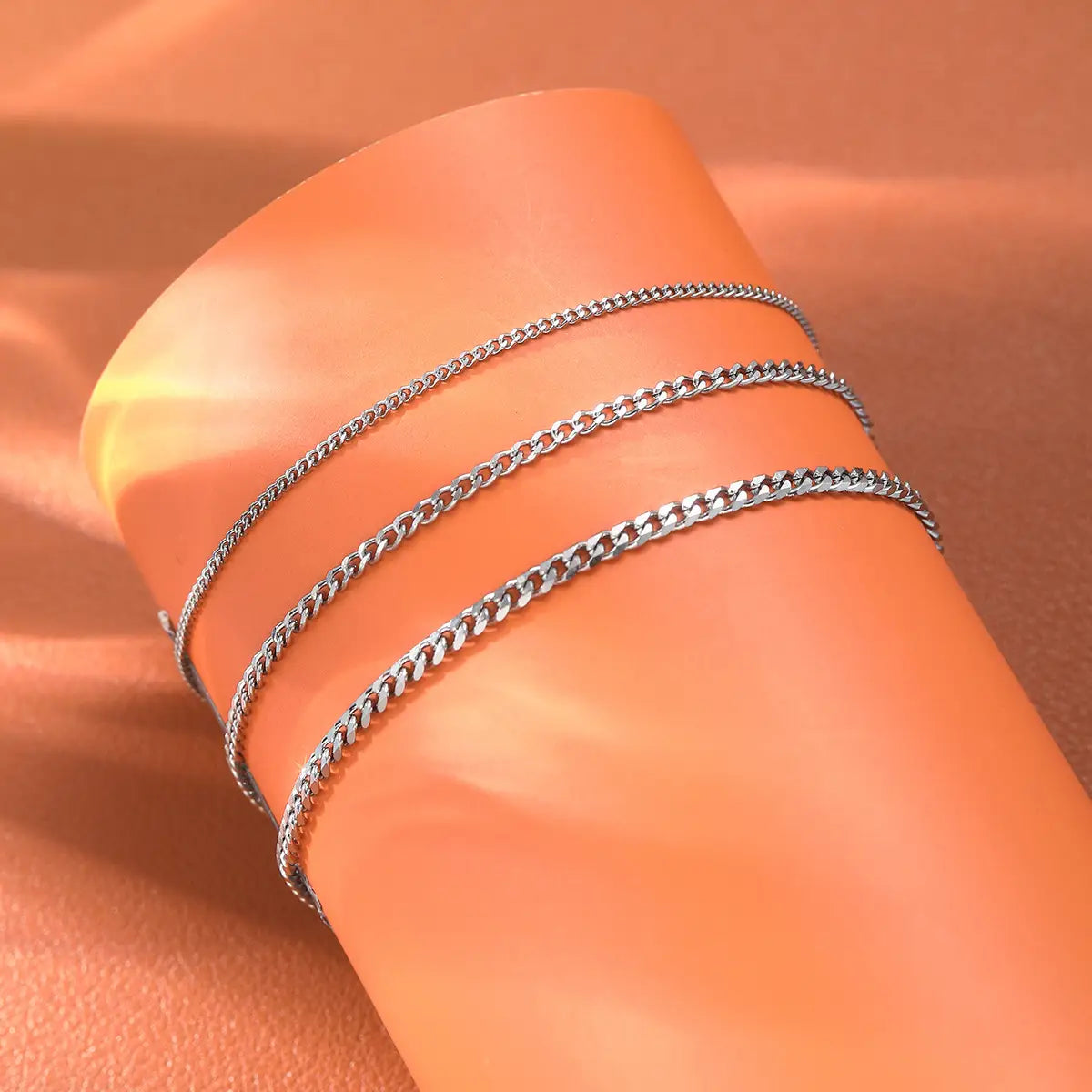 Three silver-toned curb chain anklets worn on a wrist from a unisex silver layered Cuban and Figaro link bracelet set.