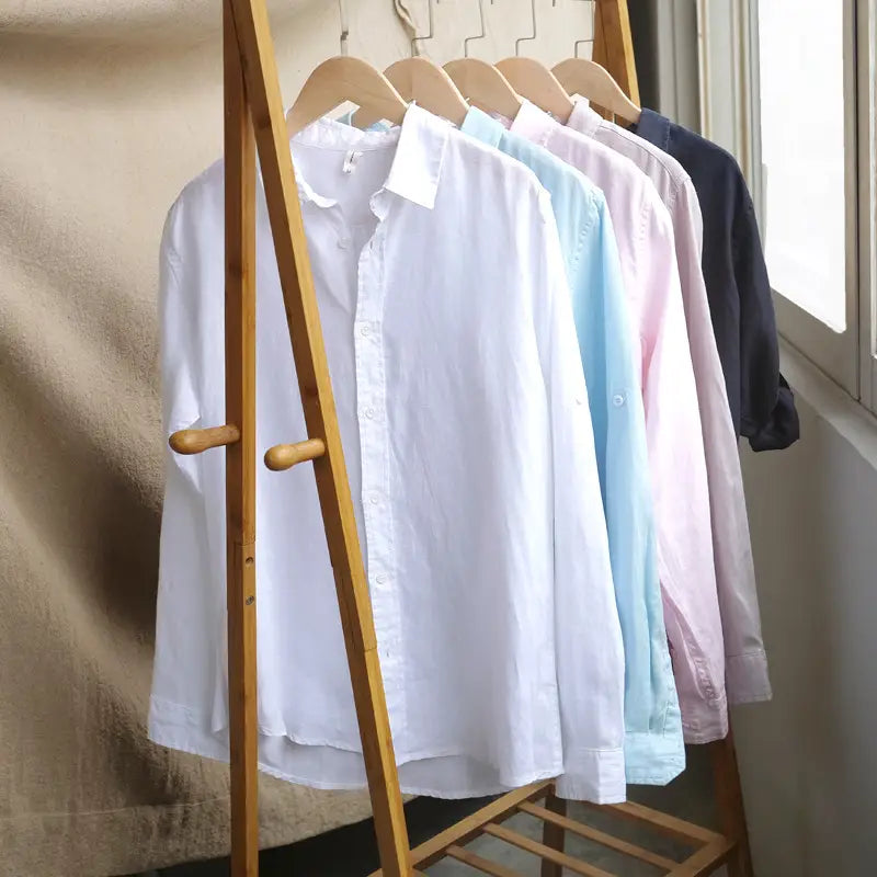 A row of men’s premium 100% linen long sleeve casual shirts in white light blue pink and navy hang neatly on wooden hangers.