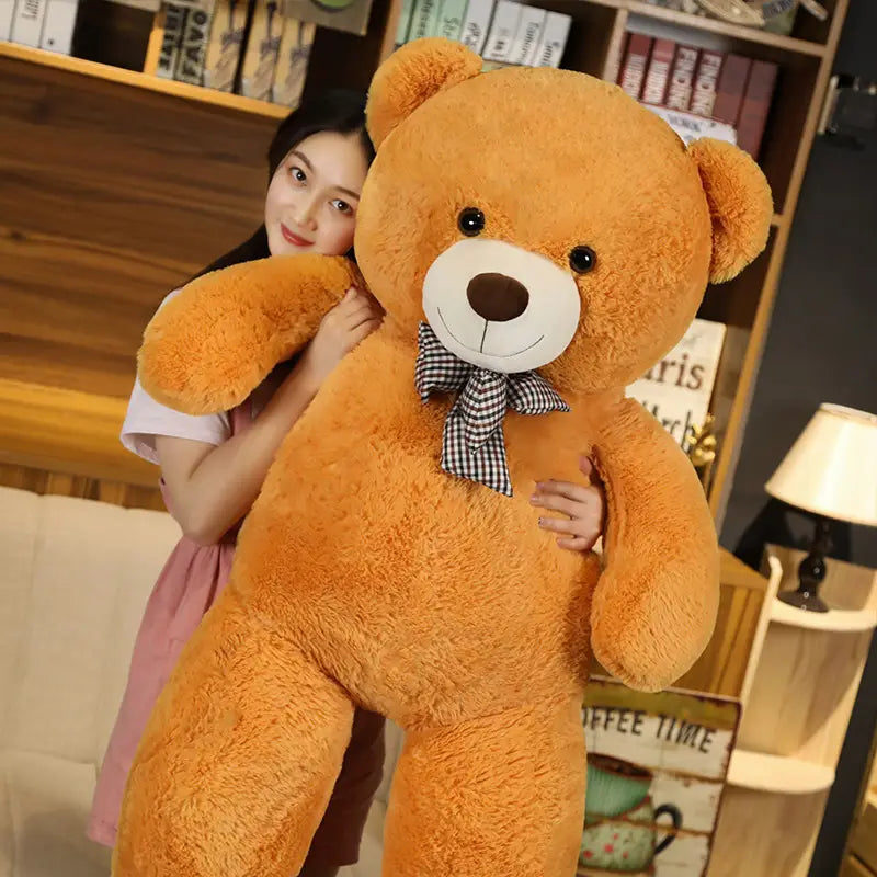 Ein flauschiger goldbrauner Teddybär mit weißem Schnäuzchen und schwarzen Knopfaugen, der eine karierte Fliege trägt, wird als Teddybär-Plüschtier mit karierter Fliege gezeigt.