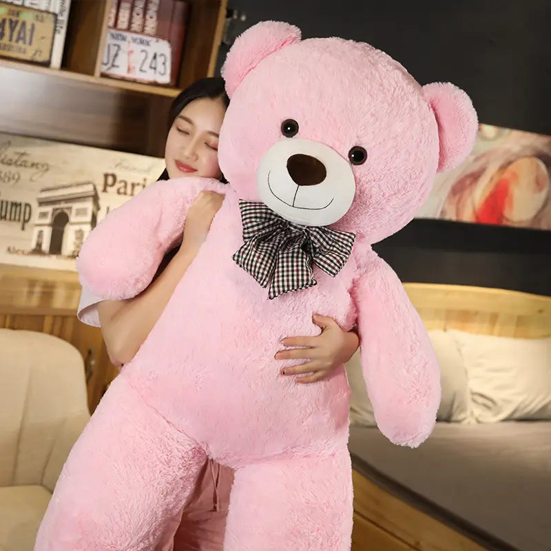 Ein flauschiger rosafarbener Teddybär mit weißem Schnäuzchen, schwarzen Knopfaugen und einer schwarz-weißen karierten Fliege, entworfen als Stofftier.