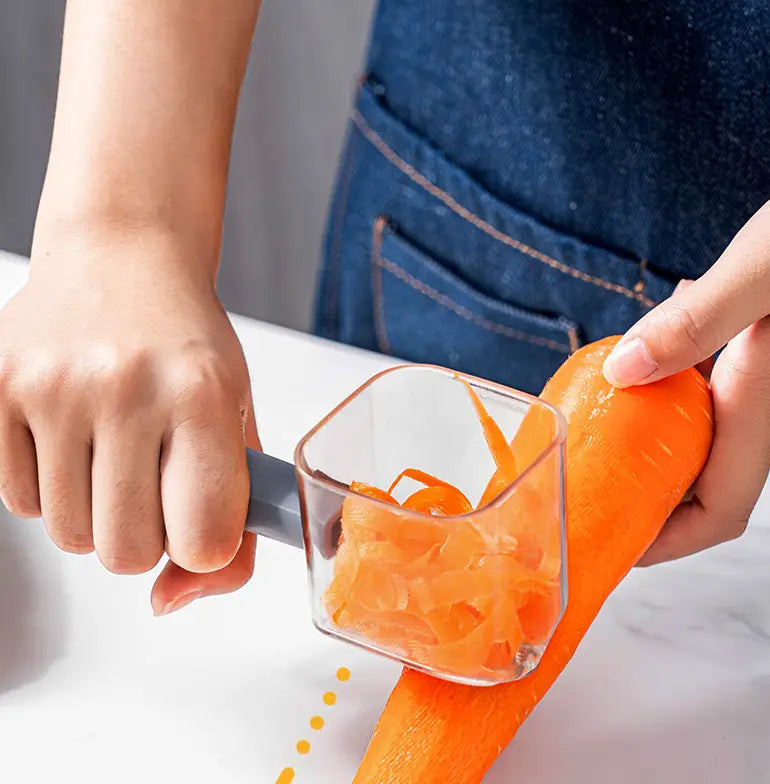 Éplucheur de légumes en acier inoxydable avec poignée grise, doté d’un protège-lame en plastique transparent conçu pour recueillir les épluchures de carotte râpées dans un compartiment carré.