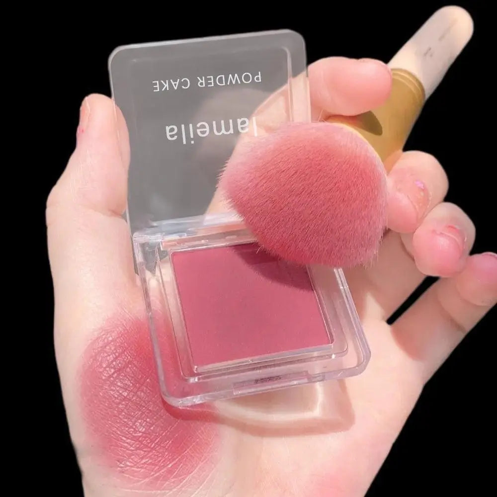 A translucent square compact of lamella powder cake blush in soft rose pink shade with a fluffy pink makeup brush from the monochrome powder blush compact with brush 4 colors collection.