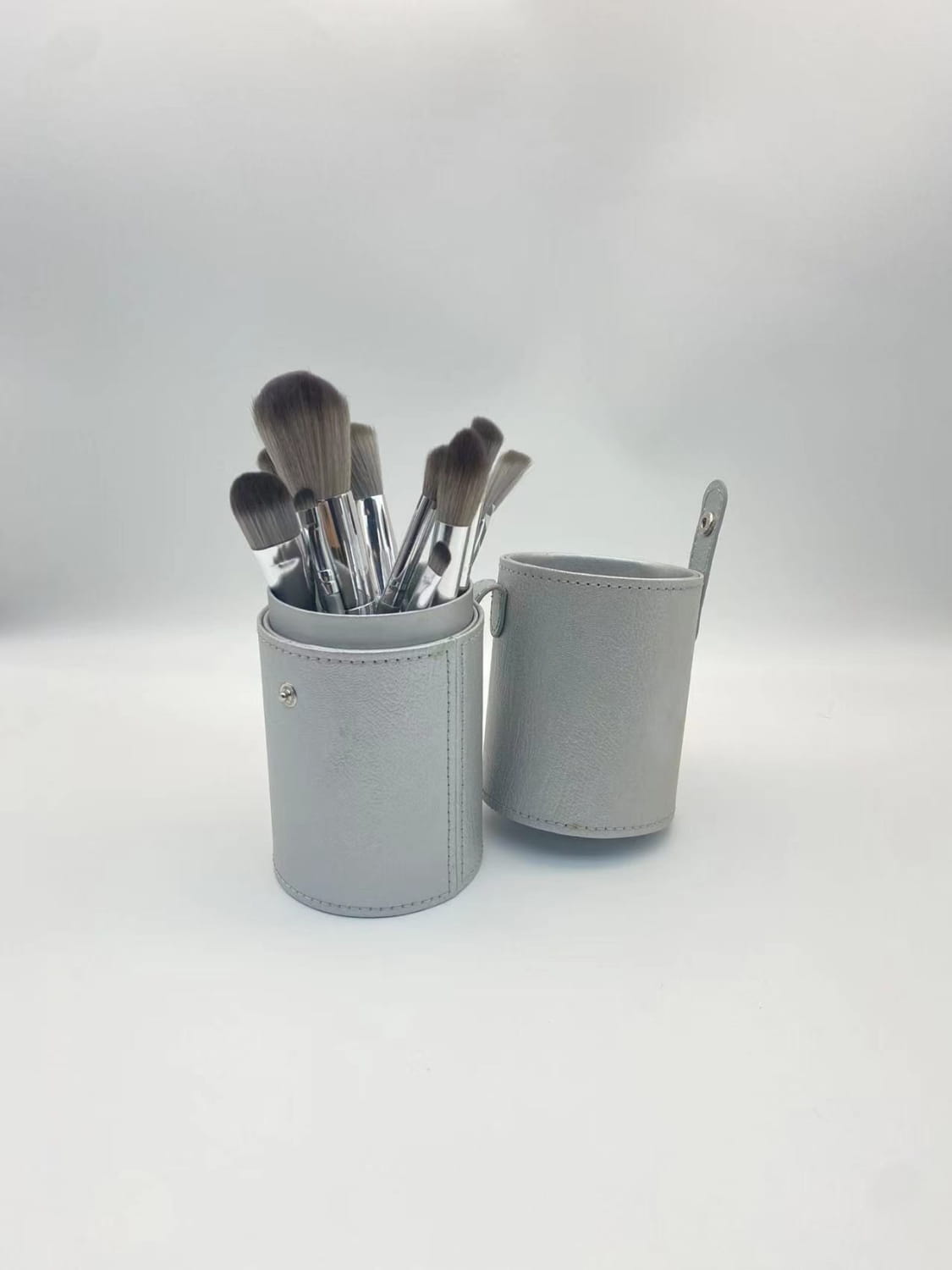 Gray leather makeup brush case storing multiple brushes from a makeup brush set with 15 silver pieces and case.