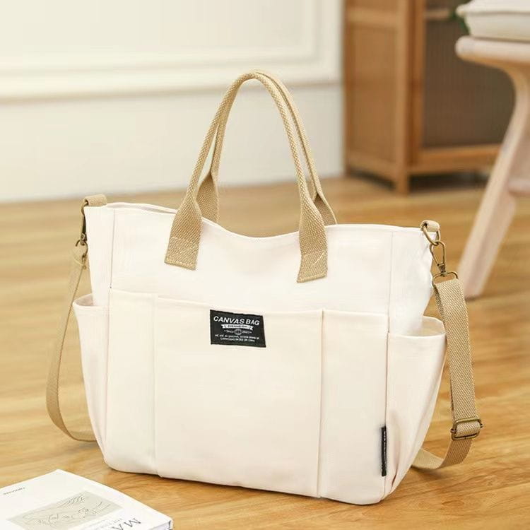 Cream-colored canvas commuter tote bag with beige leather handles and shoulder strap featuring a black label that reads canvas bag on the front.