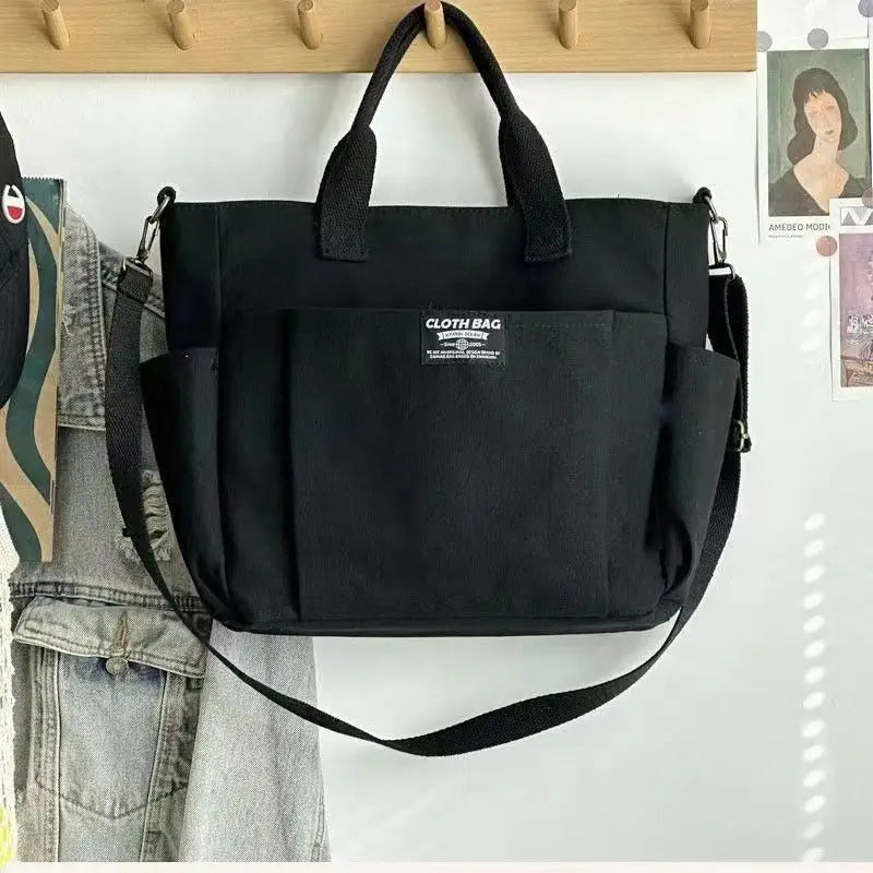 Black canvas large capacity commuter tote with sturdy leather handles and a detachable shoulder strap featuring a minimalist label patch on the front.
