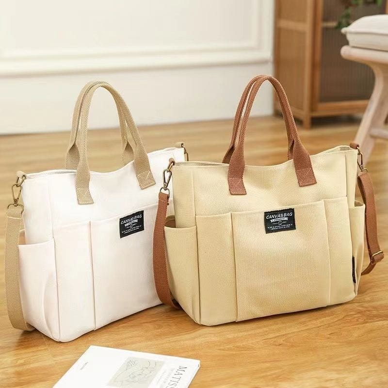 Beige large capacity canvas commuter tote with brown leather handles and matching shoulder strap featuring front pockets and black canvas bag label.