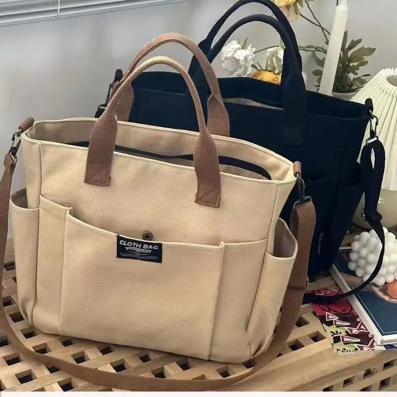 Beige canvas commuter tote bag with large capacity tan leather handles and shoulder strap featuring a front pocket with a black cloth bag label.
