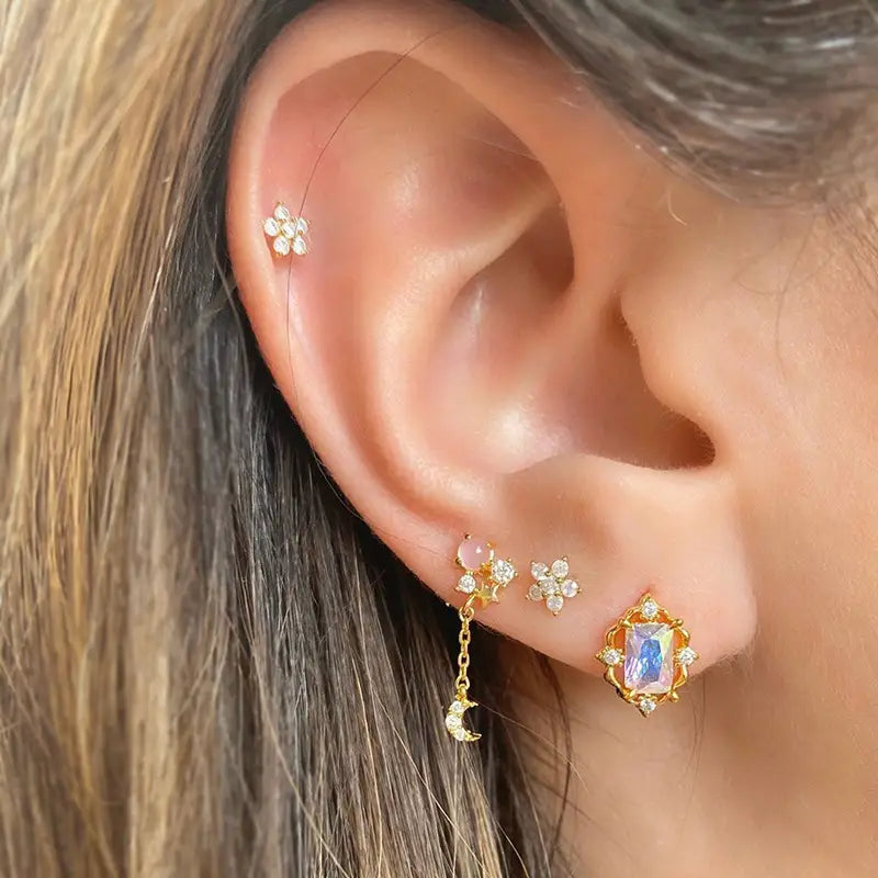 Un bijou d’oreille délicat à finition dorée, conçu avec une petite monture en forme de fleur, un pendentif suspendu en forme de lune orné de cristaux, et une boucle d’oreille rectangulaire ornée de pierres précieuses, dans l’ensemble boucles d’oreilles et manchettes « étoile et lune » en forme de papillon.