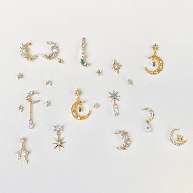 Ensemble boucles d’oreilles et manchettes « étoile et lune » en forme de papillon, comprenant des charms de nail art à thème céleste à finition dorée avec des lunes en croissant, des étoiles et des accents en forme de goutte, ornés de cristaux transparents et de pierres précieuses bleues ou vertes.
