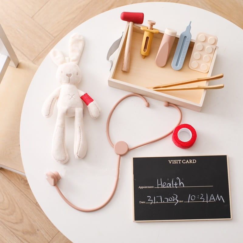 Estetoscopio de silicona color rosa pastel con auricular a juego y tubos colocados junto a un conejo de peluche blanco sobre una mesa redonda, del set Kids Doctor Kit de madera y silicona.