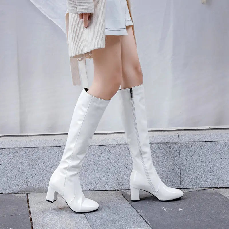 Patent leather knee high block heel square toe boots in white with a glossy finish and sturdy block heels.