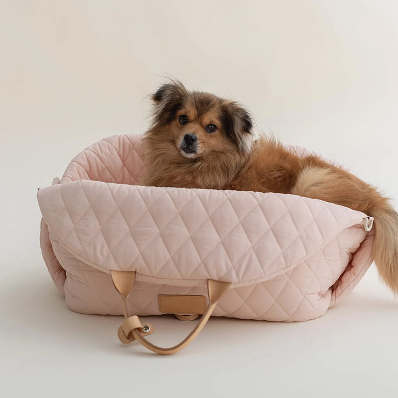 A soft blush-pink quilted travel pet carrier bag with a structured handbag-style design and tan leather handles.