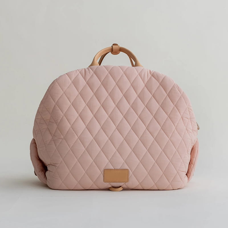 A soft blush pink quilted travel pet carrier bag with a diamond pattern tan leather handles and a matching leather plaque at the base.