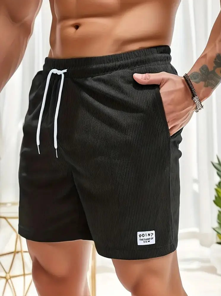 Shorts en velours côtelé noir à cordon de serrage pour hommes, avec des cordons de serrage blancs et une petite étiquette blanche sur la partie inférieure droite de la jambe, conçus pour un usage sportif.